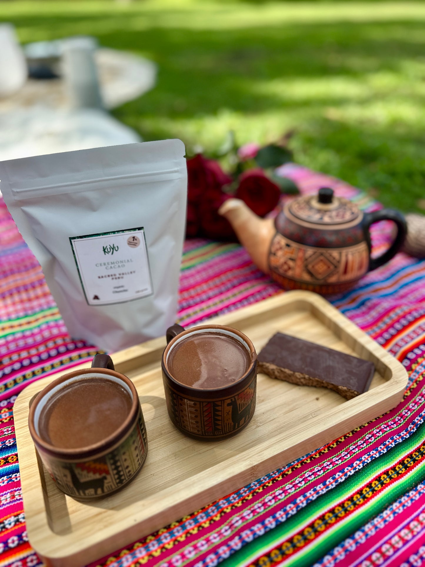 Ceremonial chocolate setup with jars, a bag, and a teapot on a colorful blanket outdoors.